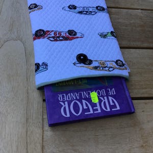 Op de afbeelding: Witte gewatteerde boekenhoes met een patroon van kleurrijke raceauto's. Een paars boek met de titel "GREGOR DE BOEKENLANDER" is gedeeltelijk zichtbaar. De hoes heeft een groene rand.