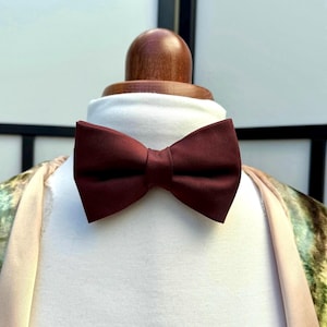 May include: A deep burgundy bow tie is presented on a white mannequin. The bow tie, made of a smooth material, is neatly tied. The mannequin has a wooden neck and is set against a neutral backdrop. A classic accessory.