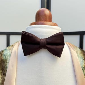 May include: A dark brown bow tie is displayed on a white shirt. The bow tie is made of a smooth fabric and is neatly tied. The shirt is on a wooden mannequin head. The background is a soft, neutral colour.