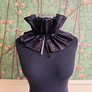 May include: A black ruffled collar made of a shiny fabric, tied with a matching ribbon. The collar is displayed on a black mannequin against a green background with a floral pattern.