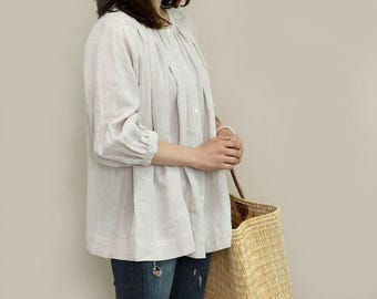 May include: A light-colored linen blouse with a round neckline and three-quarter sleeves. The blouse has a button-down front and a gathered waist. A woven straw tote bag is held in the hand.