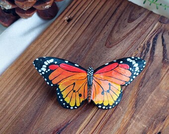 May include: A wooden butterfly brooch painted in orange, yellow, and black with white accents. The butterfly is on a wooden surface.