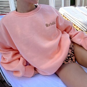 Pode incluir: Uma camisola de cor pêssego com a palavra "Marbella" bordada na frente. A camisola é usada por uma pessoa sentada numa espreguiçadeira branca.