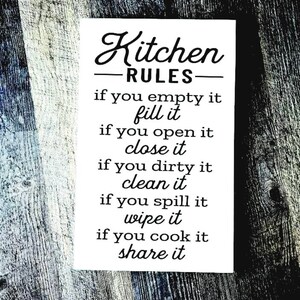 May include: A white rectangular sign with the words "Kitchen RULES" in black script. Below, a list of kitchen rules is printed in a simple, easy-to-read font. The sign is set against a rustic, wooden background.