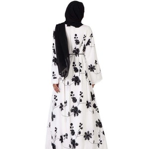 May include: A white maxi dress with black floral embroidery. The dress has long sleeves and a loose fit. The model is wearing a black headscarf.