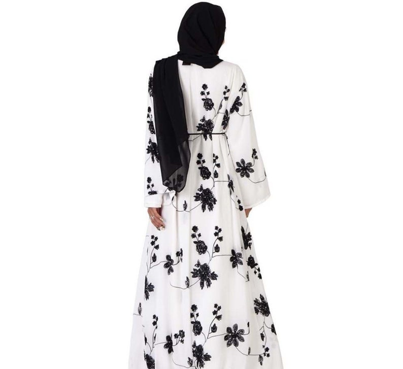 May include: A white maxi dress with black floral embroidery. The dress has long sleeves and a loose fit. The model is wearing a black headscarf.