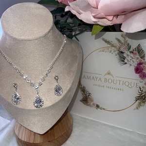May include: A silver necklace and earrings set with teardrop-shaped crystal accents. The necklace has a delicate chain and a pendant with a large crystal. The earrings are simple and elegant, with a single crystal dangling from each.