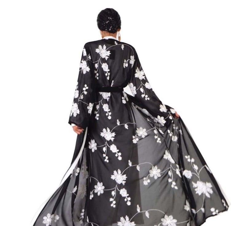 May include: A black sheer robe with white floral embroidery. The robe has a long, flowing silhouette and a sash at the waist.