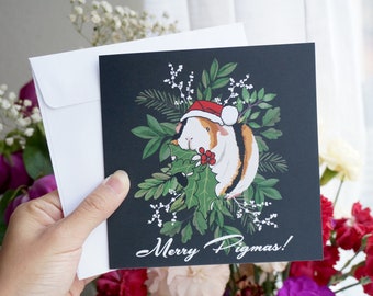 Peut inclure: Une carte de Noël représentant un cochon d'Inde portant un bonnet de Père Noël et tenant une branche de houx. La carte a un fond noir avec de la végétation verte et des fleurs blanches. Le texte "Merry Pigmas!" est imprimé en blanc au bas de la carte.