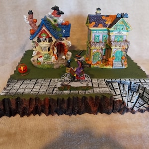 May include: A Halloween village scene featuring two decorated houses, a witch riding a bicycle, and a jack-o'-lantern. The houses are adorned with pumpkins, ghosts, and bats. The scene is set on a cobblestone base with a green lawn.