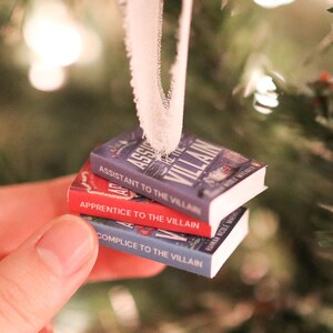 K&ouml;nnte beinhalten: Ein Stapel von drei Miniatur-Buchornamenten mit den Titeln "Assistant to the Villain", "Apprentice to the Villain" und "Complicit to the Villain". Die B&uuml;cher sind in Blau- und Rott&ouml;nen gehalten und haben wei&szlig;en Text. Ein wei&szlig;es Band ist zum Aufh&auml;ngen befestigt.