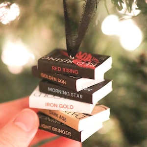 May include: A stack of miniature book ornaments with titles like "Red Rising" and "Golden Son" are suspended from a black ribbon. The books are black, white, and tan, with red and white text. The ornament hangs in front of a blurred Christmas tree.