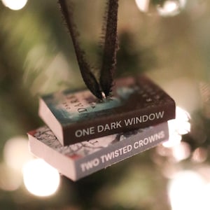 Puede incluir: Adornos de libros en miniatura, perfectos para el árbol de Navidad de un amante de los libros. Los adornos presentan los títulos "One Dark Window" y "Two Twisted Crowns" en sus portadas. Cuelgan de una cinta oscura sobre luces de árbol de Navidad.