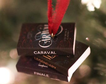 May include: Christmas tree decoration of three miniature book replicas. The books are titled "Caraval," "Legendary," and "Finale." Each book has a dark cover with gold lettering and is suspended by a red ribbon.