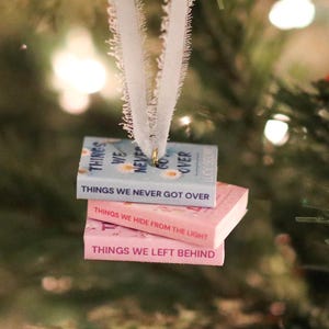 Op de afbeelding: Een kerstboomversiering met drie miniatuurboeken. De boeken zijn op elkaar gestapeld en hebben titels als "THINGS WE NEVER GOT OVER" en "THINGS WE LEFT BEHIND". De versiering hangt aan een wit lint voor een kerstboom.