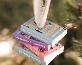May include: A stack of miniature book ornaments with titles including "Better Than The Movies", "Nothing Like The Movies", and "Betting On You". The books are in shades of blue, purple, and pink, hanging from a Christmas tree.