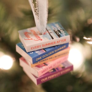 May include: A stack of miniature book ornaments with titles such as "Every Summer After" and "One Golden Summer" hangs from a white ribbon. The books are in various colors, including white, blue, and pink, against a blurred Christmas tree background.