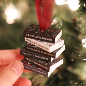 Könnte beinhalten: Ein Weihnachtsbaumschmuck mit einem Stapel Miniaturbücher, darunter Titel wie "From Blood and Ash" und "Kingdom of Flesh and Fire". Eine rote Feder ist am obersten Buch befestigt. Die Bücher werden von einem roten Band gehalten.