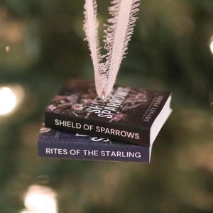 May include: A miniature book ornament featuring two stacked volumes titled Shield of Sparrows by Devney Perry and Rites of the Starling. The books are bound together with a silver eyelet and suspended by a frayed, light pink ribbon for hanging.