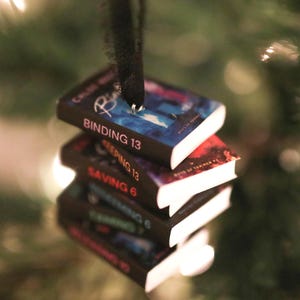 May include: A stack of miniature book ornaments, with titles such as "Binding 13" and "Keeping 13", hangs from a black ribbon. The books have colourful covers and white pages. The background is a blurred Christmas tree with lights.