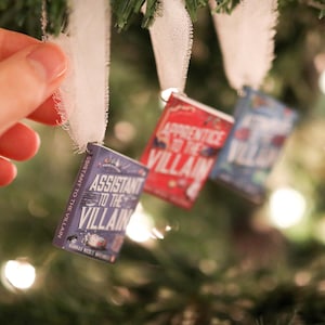 May include: Miniature book ornaments hanging on a Christmas tree. The ornaments are small, rectangular, and feature book covers with titles like "Assistant to the Villain" and "Apprentice to the Villain". Each book is attached to a white ribbon.