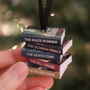 May include: A miniature book ornament stack featuring The Maze Runner, The Scorch Trials, The Death Cure, The Kill Order, and The Fever Code. The stack is suspended by a black ribbon attached to a metal eyelet at the top of the top book cover.