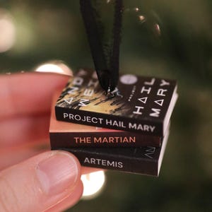 May include: A miniature ornament featuring a stack of three books by Andy Weir, including Project Hail Mary, The Martian, and Artemis. The books are bound together with a black ribbon loop attached to the top for hanging as a decorative item.