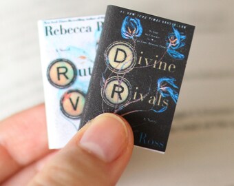 May include: Two miniature books are held in a hand. One book has the title "Rebecca R" and "Rut V" on the cover. The other book is black with the title "Divine Rivals" and floral accents. The books are small, likely for a dollhouse or craft project.