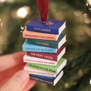 May include: A miniature stack of books ornament featuring titles from the Outlander series including Dragonfly in Amber, Voyager, Drums of Autumn, The Fiery Cross, A Breath of Snow and Ashes, An Echo in the Bone, and Written in My Own Heart's Blood.