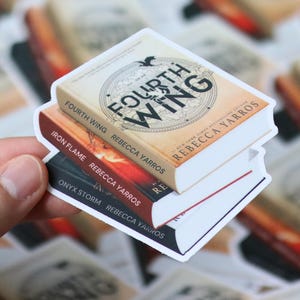 Pode incluir: Pilha de três autocolantes de livros com os títulos "Fourth Wing", "Iron Flame" e "Onyx Storm" de Rebecca Yarros. O autocolante superior tem uma capa bege com um design circular e o título "Fourth Wing". Os outros dois autocolantes têm capas vermelhas e pretas.
