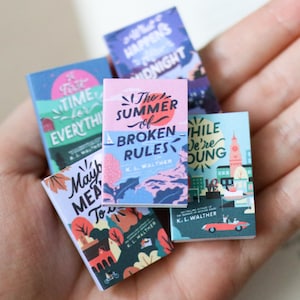 May include: A selection of miniature books with vibrant covers, held in a hand. Titles include "The Summer of Broken Rules" and "While We're Young." Each book displays unique illustrations and text.