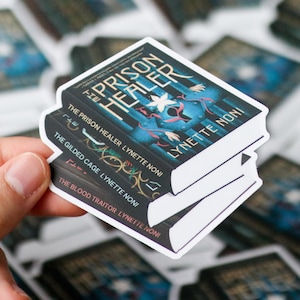 May include: A stack of three book stickers with white borders. The top sticker reads "The Prison Healer" by Lynette Noni, featuring a blue and teal cover design. The other stickers display "The Gilded Cage" and "The Blood Traitor" by the same author.