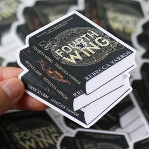 Puede incluir: Una pila de tres pegatinas con forma de libro. La pegatina superior dice "Fourth Wing" en letras doradas. Las otras pegatinas dicen "Iron Flame" y "Onyx Storm" de Rebecca Yarros. Las pegatinas son negras con bordes blancos.