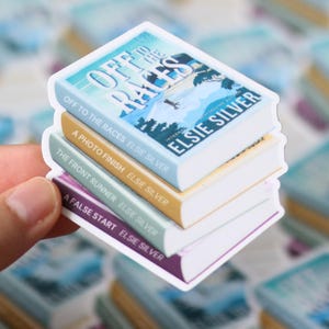 May include: A stack of four book stickers with the titles "Off to the Races," "A Photo Finish," "The Front Runner," and "A False Start" by Elsie Silver. The top sticker has a blue cover with a landscape design. The other stickers have tan, green, and purple covers.