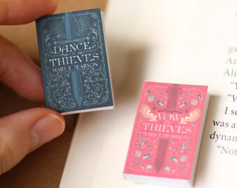May include: Two miniature books with the titles "Dance of Thieves" and "Vow of Thieves" by Mary E. Pearson. The first book has a blue cover, and the second has a pink cover. Both books have decorative designs and a sword illustration.