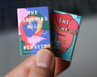 May include: Two miniature books with colorful covers. The front book is titled The Vanishing Half by Brit Bennett, featuring abstract shapes in pink, blue, and green. The partially visible book behind it is titled The Mothers by Brit Bennett.