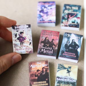 Puede incluir: Conjunto de libros en miniatura de Brandon Sanderson, con títulos como "The Hero of Ages" y "The Lost Metal". Las portadas de los libros muestran ilustraciones y texto vibrantes. Estos pequeños libros son probablemente para una casa de muñecas o un proyecto de manualidades.