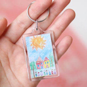 Peut inclure: Un porte-cl&eacute;s en plastique transparent avec un dessin color&eacute; d'un soleil, des maisons et des personnes &agrave; l'int&eacute;rieur. Le dessin est prot&eacute;g&eacute; par une couverture en plastique transparent.