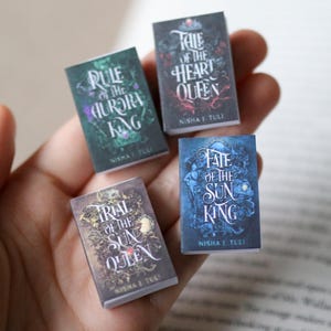 Puede incluir: Cuatro libros de fantasía en miniatura con títulos como "Rule of the Aurora King" y "Tale of the Heart Queen." Cada libro tiene un diseño de portada único con ilustraciones intrincadas y el nombre del autor, Nisha I. Tuli.