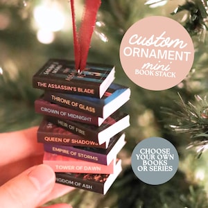 以下が含まれることがあります： 「The Assassin's Blade」や「Throne of Glass」などのタイトルが書かれたミニチュア本のスタックオーナメント。赤いリボンが付いています。画像には「custom ornament mini book stack」と「choose your own books or series」という文字が書かれています。