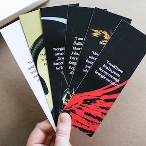May include: A collection of rectangular bookmarks with quotes printed on them. The bookmarks have a black background with white text. Some bookmarks have red and yellow graphic designs. The bookmarks are held in a hand.