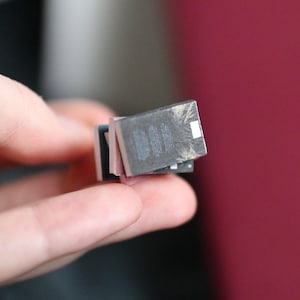 Puede incluir: Una pila de libros en miniatura, sostenidos en una mano. Los libros tienen cubiertas de color gris oscuro con detalles blancos y bordes rosas y azules. El fondo es una mezcla borrosa de rojo y gris.
