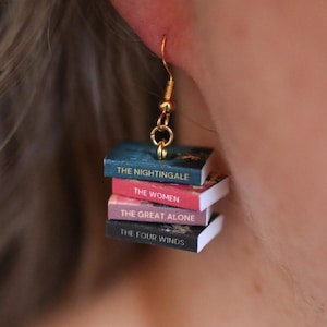 Puede incluir: Pendientes colgantes dorados con pilas de libros en miniatura. Cada libro tiene un t&iacute;tulo diferente: "The Nightingale", "The Women", "The Great Alone" y "The Four Winds". Un accesorio &uacute;nico para los amantes de la lectura.