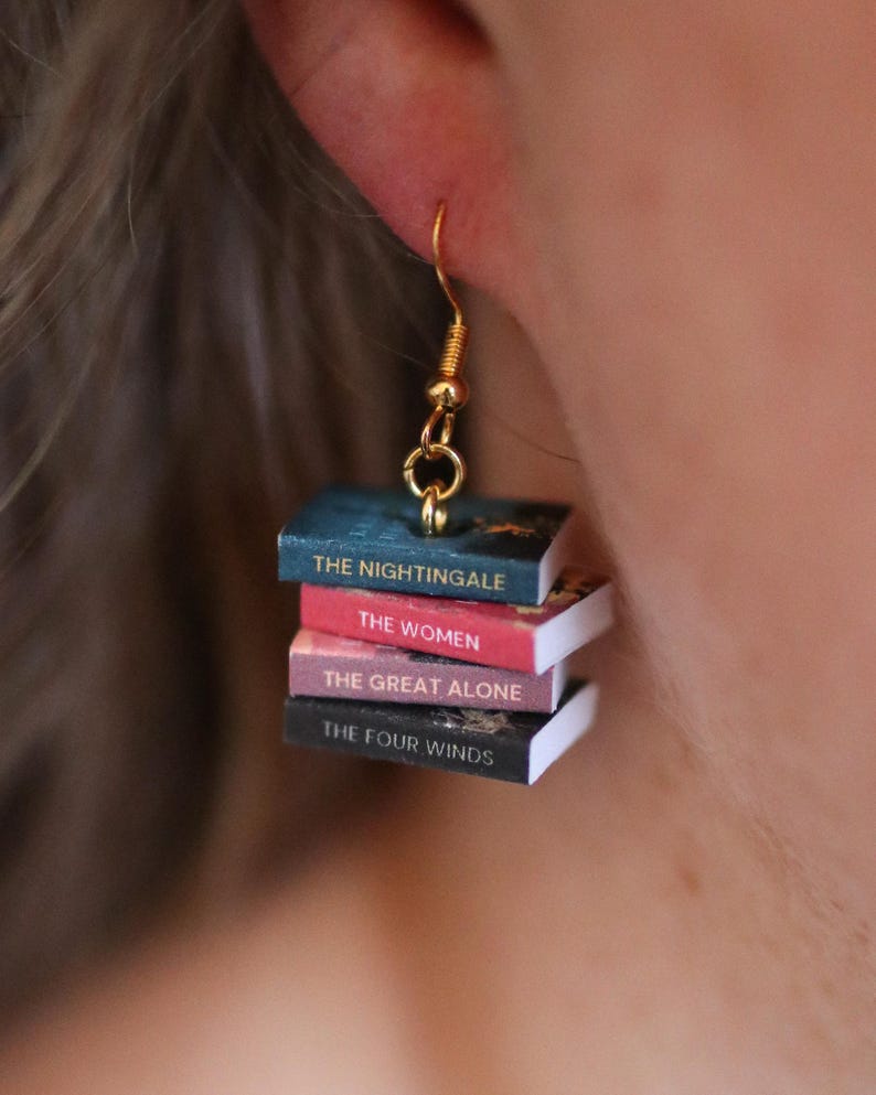 Puede incluir: Pendientes colgantes dorados con pilas de libros en miniatura. Cada libro tiene un t&iacute;tulo diferente: "The Nightingale", "The Women", "The Great Alone" y "The Four Winds". Un accesorio &uacute;nico para los amantes de la lectura.