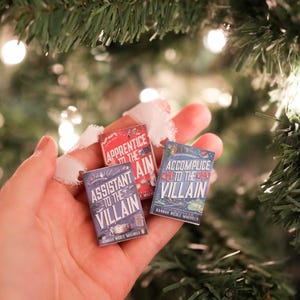 May include: Three miniature book ornaments with titles "Apprentice to the Villain", "Assistant to the Villain", and "Accomplice to the Villain". The books are held in a hand, with a Christmas tree in the background. The books are red and blue.