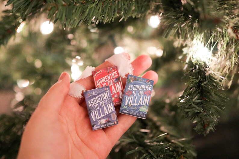 May include: Three miniature book ornaments with titles "Apprentice to the Villain", "Assistant to the Villain", and "Accomplice to the Villain". The books are held in a hand, with a Christmas tree in the background. The books are red and blue.