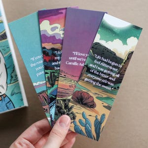 Puede incluir: Una mano sostiene cuatro marcapáginas coloridos con ilustraciones de paisajes desérticos y citas. Los marcapáginas presentan tonos de azul, morado y naranja. Cada marcapáginas tiene una cita diferente en texto blanco. Las citas son: "Si haces el camino que pavimentas", "Yo sigo mi y t atrás", "Te amaré hasta que seamos Camille Ash", y "La vida había comenzado a sentirse agridulce, y yo estaba recibiendo todo lo amargo y todos los demás estaban recibiendo lo dulce."