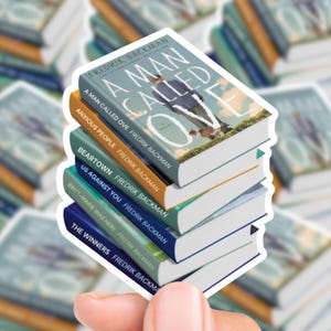 May include: A stack of stickers featuring book covers by Fredrik Backman. Titles include "A Man Called Ove," "Anxious People," and "Beartown." The stickers have white borders and are held by a hand, with other book stickers in the background.