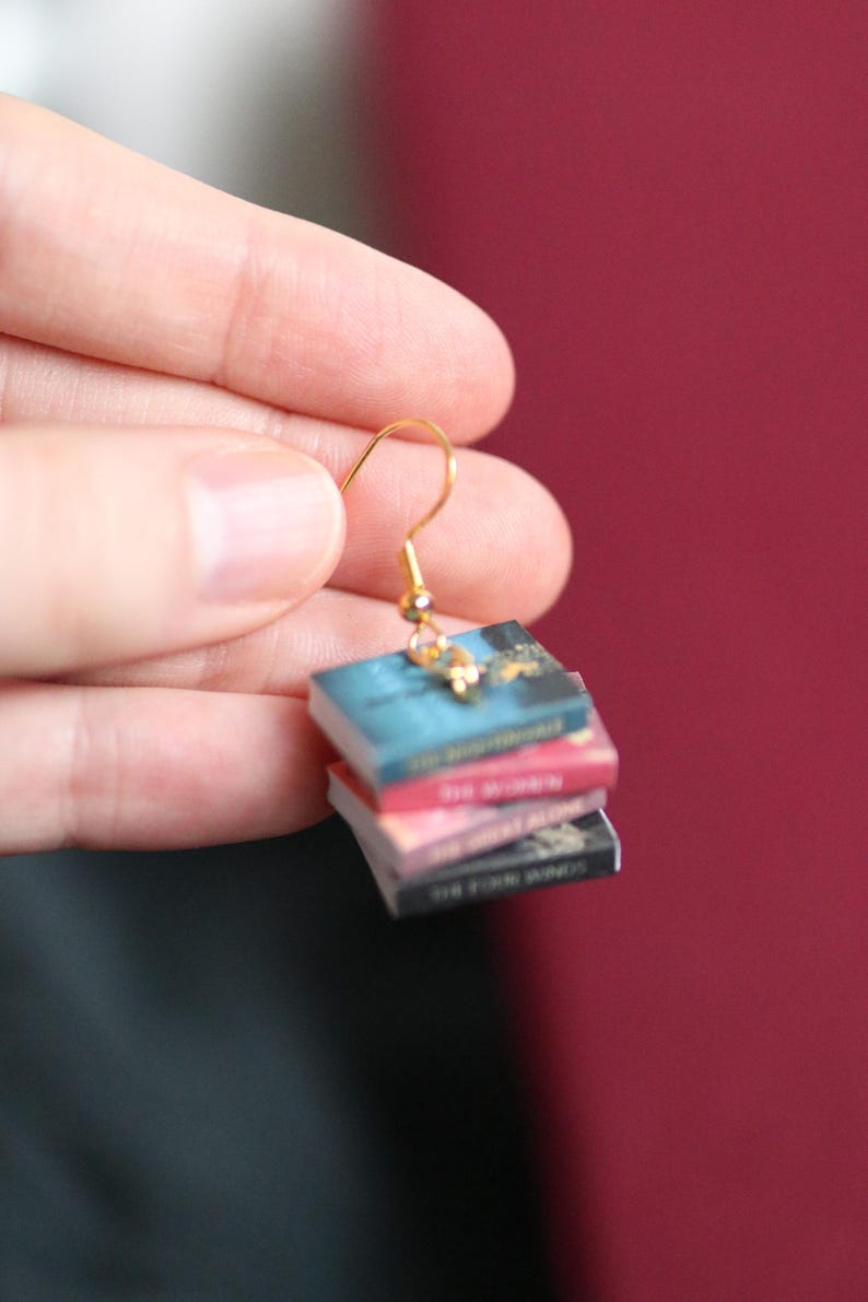 Puede incluir: Un pendiente dorado con un dise&ntilde;o de libros en miniatura apilados. Los libros tienen cubiertas azules, rosas y negras, con detalles dorados. El pendiente est&aacute; suspendido de un gancho dorado y una peque&ntilde;a cuenta dorada.