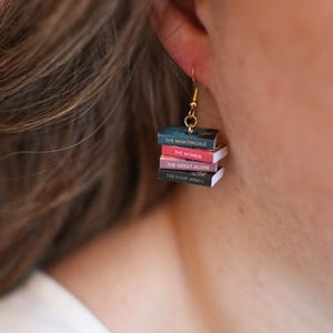 Puede incluir: Pendientes colgantes dorados con mini pilas de libros. Cada libro tiene un t&iacute;tulo diferente: "The Nightingale", "The Women", "The Great Alone" y "The Four Winds". Un accesorio &uacute;nico para los amantes de los libros.
